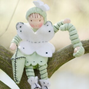 Puede incluir: Una mu&ntilde;eca de fieltro verde con alas de mariposa blancas y una oruga de fieltro verde. La mu&ntilde;eca est&aacute; sentada en una rama.