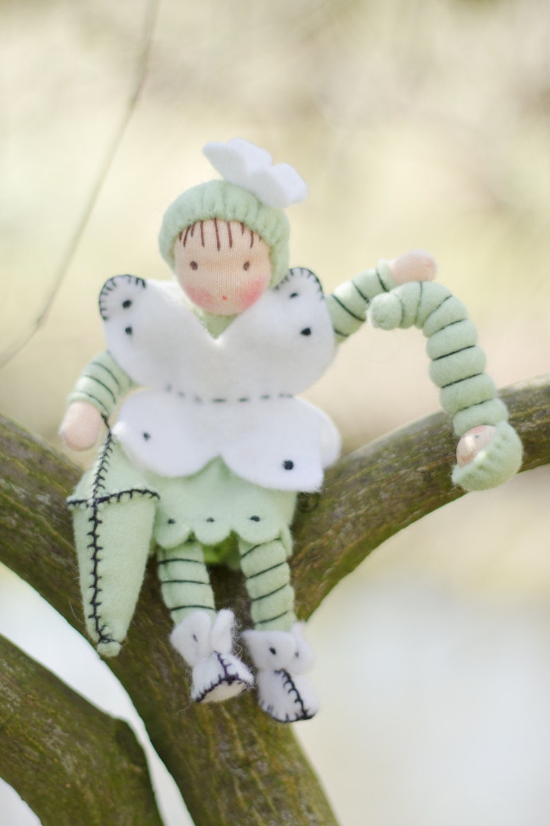 Puede incluir: Una mu&ntilde;eca de fieltro verde con alas de mariposa blancas y una oruga de fieltro verde. La mu&ntilde;eca est&aacute; sentada en una rama.