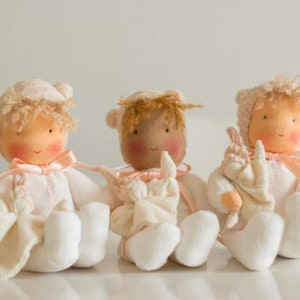 May include: Three handmade cloth dolls with pink and white clothing and rosy cheeks. The dolls are holding small white cloth animals.