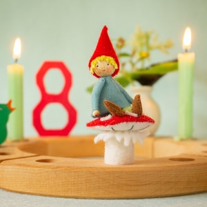 Puede incluir: Un anillo de velas de cumpleaños de madera con un número ocho rojo, dos velas verdes y un gnomo de fieltro sentado en un hongo rojo. El gnomo lleva una camisa azul y un gorro rojo.
