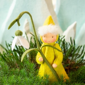 Op de afbeelding: Een gele viltpop met een witte viltmuts en een witte viltbaard houdt een groene viltsteel vast met een witte viltbloem. De pop zit in een bed van groene vilt.