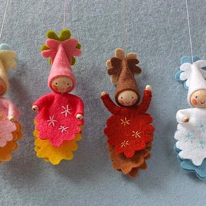 Puede incluir: Cuatro muñecas de fieltro hechas a mano con cabezas de madera y caras sonrientes. Cada muñeca tiene una combinación de colores única: rosa, rojo, naranja y blanco. Están decoradas con estrellas bordadas y faldas de fieltro en capas, colgadas sobre un fondo azul claro.