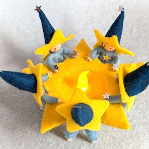 Op de afbeelding: Een handgemaakte vilten sterdecoratie met twee kleine figuren. De ster is geel met blauwe accenten en een centraal licht. De figuren dragen gele sterhoeden en blauwe kleding, en houden stervormige ornamenten vast. Een charmant, handgemaakt item.