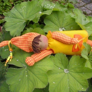 Puede incluir: Una muñeca hecha a mano con un vestido a cuadros amarillo y naranja, cabello castaño y rostro rosado. La muñeca está tumbada sobre un lecho de hojas verdes.