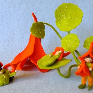 May include: Four felt dolls dressed in orange and green, sitting on a felt flower with green leaves. The dolls are handmade and have a whimsical, fairy-like appearance.