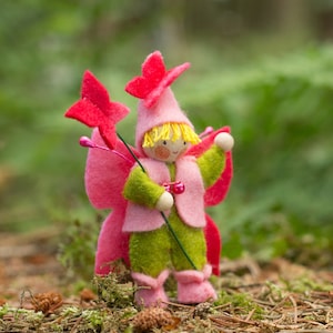 May include: A felt doll wearing a pink dress with green accents and a flower crown. The doll is holding a pink flower in its hand.