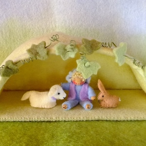 May include: A felted nativity scene featuring a cream-colored shelter with green leaf and wire accents. Inside are felt figures of a lamb, a seated figure in blue and purple, and a brown rabbit. The scene is set on a green surface.