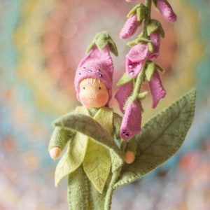 May include: A handmade felt doll dressed as a fairy, holding a large leaf and a stem of pink foxglove flowers. The doll wears a green outfit with leaf accents, a pink hat, and pink shoes. The background is a soft, blurred mix of colors.