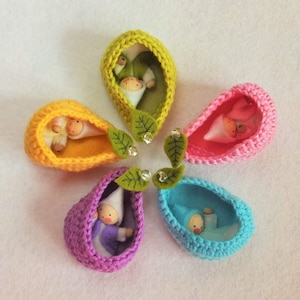 May include: Six crocheted doll cradles in different colors, each containing a small felt doll with a white bonnet. The cradles are arranged in a circle with green felt leaves and silver bells.
