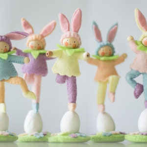 May include: Five felt bunny figurines stand on white eggs. Each bunny is wearing a different colour outfit: light blue, lavender, yellow, orange, and pink. The bunnies are all balancing on one leg and have their arms outstretched.