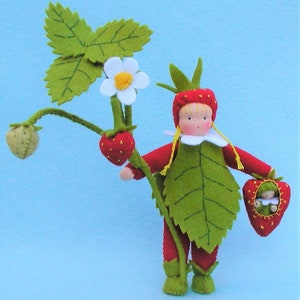 May include: A felt doll dressed as a strawberry with a green felt leaf costume, holding a smaller felt strawberry doll. The doll is standing next to a felt strawberry plant with a white flower.