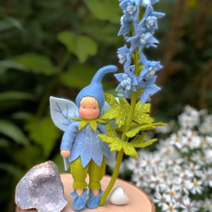 Puede incluir: Una muñeca de hadas hecha a mano con ropa azul y pantalones verdes está junto a una flor azul de fieltro. La muñeca tiene una cara amarilla y alas. Una geoda, una piedra blanca y una pluma descansan sobre una rodaja de madera.