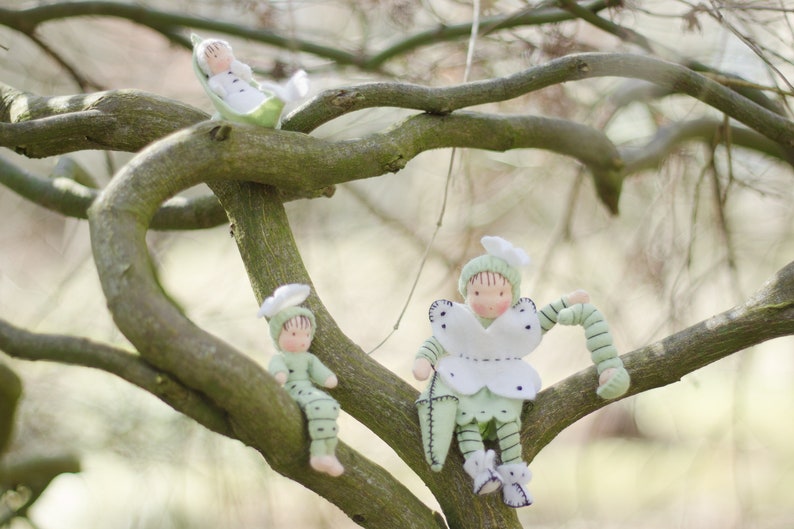 Puede incluir: Tres mu&ntilde;ecas de fieltro hechas a mano vestidas con atuendos verdes y blancos est&aacute;n posadas en ramas de &aacute;rboles. Las mu&ntilde;ecas tienen caras pintadas y llevan sombreros.