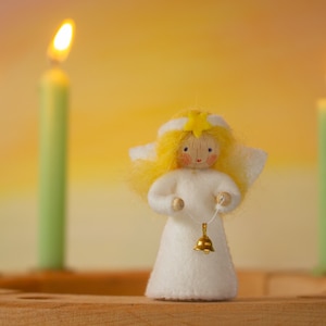 May include: A white felt angel doll with blonde hair and a yellow star on its head. The angel is holding a gold bell on a string. The angel is standing in front of three green candles.