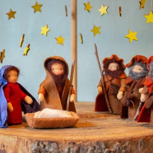 May include: A nativity scene with three wise men, a mother and child, and a manger. The figures are made of felt and have a rustic look. The scene is set against a blue background with gold stars.