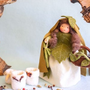 Puede incluir: Una muñeca de fieltro con un vestido de hojas verdes y cabello castaño, sosteniendo una pequeña cesta marrón con hojas verdes. La muñeca está de pie frente a un fondo de madera.