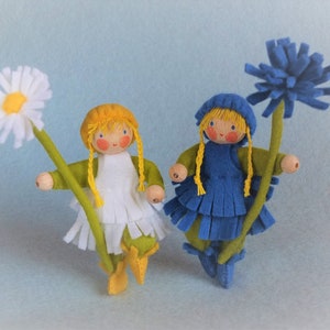 May include: Two felt dolls with yarn hair holding flowers. The doll on the left is wearing a white dress and green skirt and holding a white daisy. The doll on the right is wearing a blue dress and holding a blue flower.