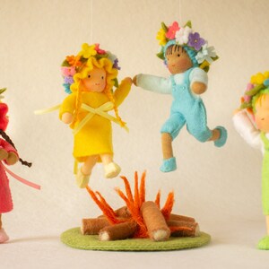 Pode incluir: Quatro bonecas de feltro com coroas de flores pulam em torno de uma pequena fogueira de feltro. As bonecas usam roupas coloridas e têm rostos pintados.