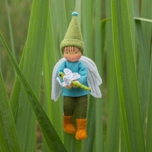 Op de afbeelding: Een vilten pop met een groene hoed en een blauw shirt die een groene libel vasthoudt. De pop heeft witte vleugels en staat in een veld met groen gras.