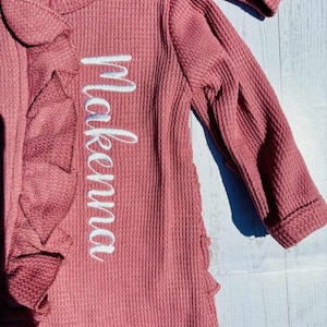 Puede incluir: Conjunto de beb&eacute; de punto de gofre color rosa empolvado con el nombre "Makena" bordado en escritura blanca. Presenta un ribete con volantes y mangas largas. La tela tiene un aspecto texturizado.