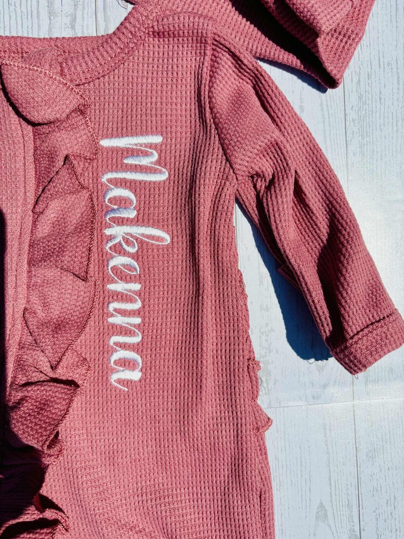 Puede incluir: Conjunto de beb&eacute; de punto de gofre color rosa empolvado con el nombre "Makena" bordado en escritura blanca. Presenta un ribete con volantes y mangas largas. La tela tiene un aspecto texturizado.
