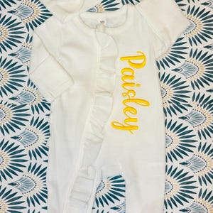 Puede incluir: Un pelele blanco para beb&eacute; con el nombre "Paisley" bordado en amarillo. El pelele tiene un ribete con volantes y se combina con una diadema blanca con lazo a juego. El conjunto se muestra sobre un fondo de tela estampada.