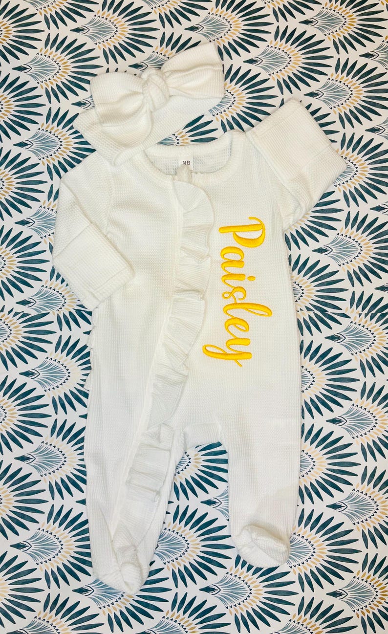 Puede incluir: Un pelele blanco para beb&eacute; con el nombre "Paisley" bordado en amarillo. El pelele tiene un ribete con volantes y se combina con una diadema blanca con lazo a juego. El conjunto se muestra sobre un fondo de tela estampada.