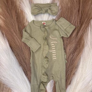 Puede incluir: Conjunto de ropa de beb&eacute; verde oliva con una diadema a juego. El mono de manga larga presenta una textura de punto de gofre, ribete de volantes y el nombre "Mayna" bordado. La diadema tiene un detalle de lazo.