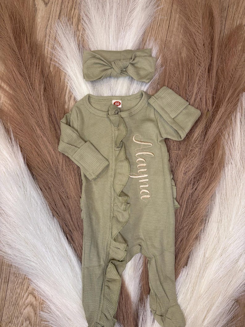 Puede incluir: Conjunto de ropa de beb&eacute; verde oliva con una diadema a juego. El mono de manga larga presenta una textura de punto de gofre, ribete de volantes y el nombre "Mayna" bordado. La diadema tiene un detalle de lazo.