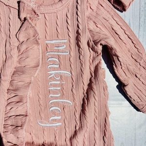 Puede incluir: Un conjunto rosa para reci&eacute;n nacido con dise&ntilde;o de punto trenzado. El conjunto tiene mangas largas, cremallera y detalle de volantes. El nombre "Makinsley" est&aacute; bordado en blanco. La palabra "newborn" est&aacute; en una etiqueta.