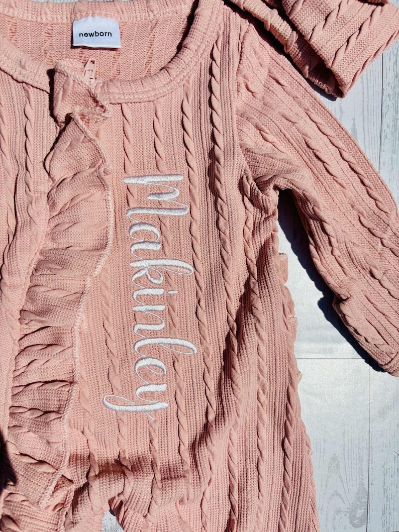 Puede incluir: Un conjunto rosa para reci&eacute;n nacido con dise&ntilde;o de punto trenzado. El conjunto tiene mangas largas, cremallera y detalle de volantes. El nombre "Makinsley" est&aacute; bordado en blanco. La palabra "newborn" est&aacute; en una etiqueta.