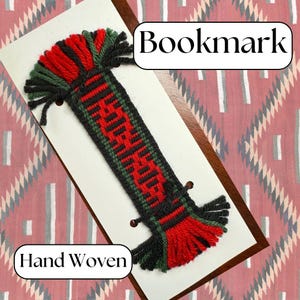 May include: Hand-woven bookmark with a red, green, and black design. The bookmark has a rectangular shape with a woven pattern in the center and tassels at both ends. The word "Bookmark" is in a white box at the top.