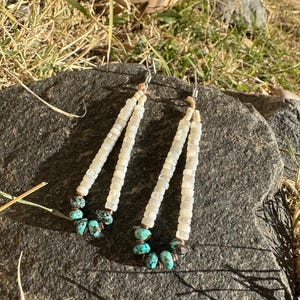 May include: A pair of handmade earrings featuring white, rectangular beads strung together in a cascading design. The earrings have turquoise-colored stone accents at the bottom and are suspended from silver-colored hooks. The earrings are displayed on a dark gray stone.