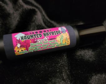 May include: A pink and black roll-on bottle with a label that says "Haunted Hayride" and lists the scent notes: hazelnut, hot cocoa, cinnamon sugar donuts, maple, hay bales, and creaky old-wood.