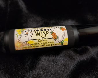 Può includere: Un flacone roll-on nero e giallo con un design a forma di fantasma e fiori. L'etichetta recita "Lovesick Witchery WISP Lemon, vanilla, lemon curd, marshmallow fluff, and pie crust."