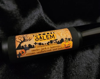 May include: A black and orange roll-on bottle with a Halloween-themed label. The label features a spooky cityscape with a pumpkin, spiderwebs, and the text "LOVESICK WITCHERY SALEM Cinnamon, leaves, smoke, jack o' lanterns, creamy sandalwood, and amber."
