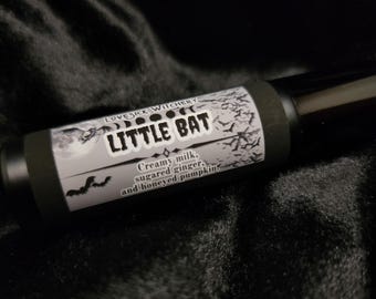 Può includere: Un flacone nero roll-on con un'etichetta bianca che dice "Lovesick Witchery Little Bat Creamy milk, sugared ginger, and honeyed pumpkins".