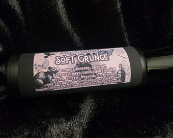May include: A black cylindrical product with a pink label featuring the text "SOFT GRUNGE" and a list of scent notes. The label includes illustrations of a skull, a rat, and a bottle. The product is set against a dark, textured background.