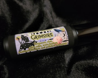 May include: A black roller bottle with a purple and yellow label that reads "Lovesick Witchery Grimoire Oil". The label also lists the scent notes: Bergamot, chai, spice cake, worn leather, cashmere blanket, and polished mahogany.