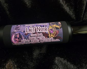 May include: A black bottle with a label featuring the text "LATT NOIR" and the words "LOVESICK WITCHERY". The label includes illustrations of a skull, butterfly, and a plant with a purple flower. The label also lists the ingredients: clove bud, heavy cream, buttery vanilla, black musk, smoldering patchouli, and black vanilla.