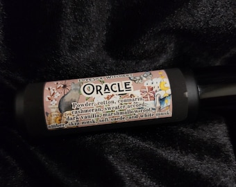 May include: A black cylindrical perfume bottle with the word "ORACLE" in white text. The label includes the text "LOVESICK WITCHERY" and lists fragrance notes such as powder, cotton, and amber. The bottle rests on a dark, textured surface.