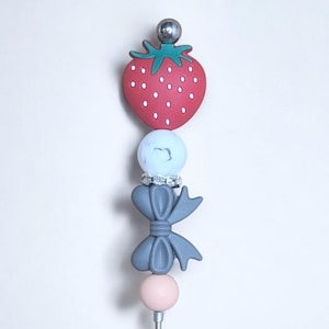 May include: A red strawberry charm with a green stem and leaves, a white bead with a silver ring, a gray bow, and a pink bead on a silver metal skewer.