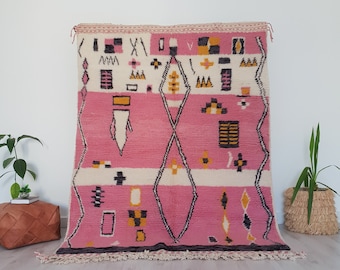 May include: A pink and white Moroccan rug with geometric patterns in black, yellow, and orange. The rug has a fringe border.