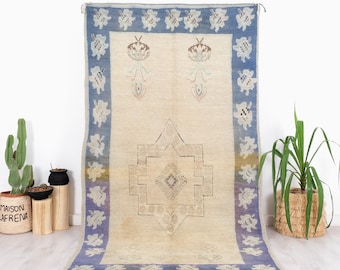 May include: A vintage rug with a cream background and a blue border. The rug has a geometric pattern in the center and floral motifs in the corners.
