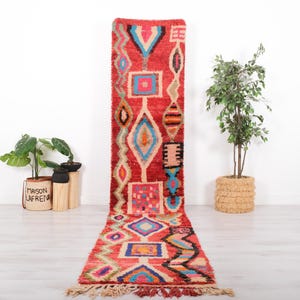 May include: A long, narrow runner rug featuring a vibrant red background with a repeating pattern of geometric shapes including diamonds, squares, and zigzags in bright shades of blue, yellow, pink, orange, and black, finished with fringe.