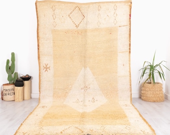 May include: A large, rectangular rug with a cream and tan color scheme. The rug features geometric patterns, including diamonds and triangles, and has a textured appearance. The edges are bordered with a darker tan color.