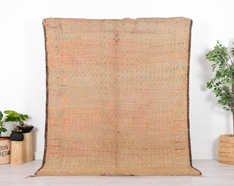 May include: A large, rectangular rug with a beige and orange geometric pattern. The rug has a dark brown border and fringe along the bottom edge. The rug is displayed against a white wall.