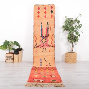 May include: A long, rectangular runner rug featuring a gradient from light peach to vibrant red. The rug displays geometric patterns, including diamonds and abstract shapes in black, blue, pink, and white, finished with a fringed edge at the bottom.