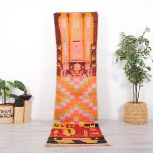 Può includere: Un tappeto passatoia lungo e stretto caratterizzato da un vivace motivo geometrico multicolore. Il design include sezioni rosa, arancioni, beige e rosso scuro, con motivi astratti e una sezione a scacchi al centro, rifinito con frange.