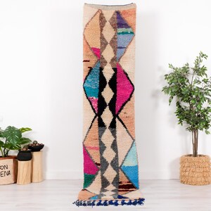 May include: A long, rectangular rug with a geometric diamond pattern in shades of beige, pink, black, blue, and gray. The rug has a textured, woven appearance and a dark blue fringe along the bottom edge. The rug is displayed vertically.
